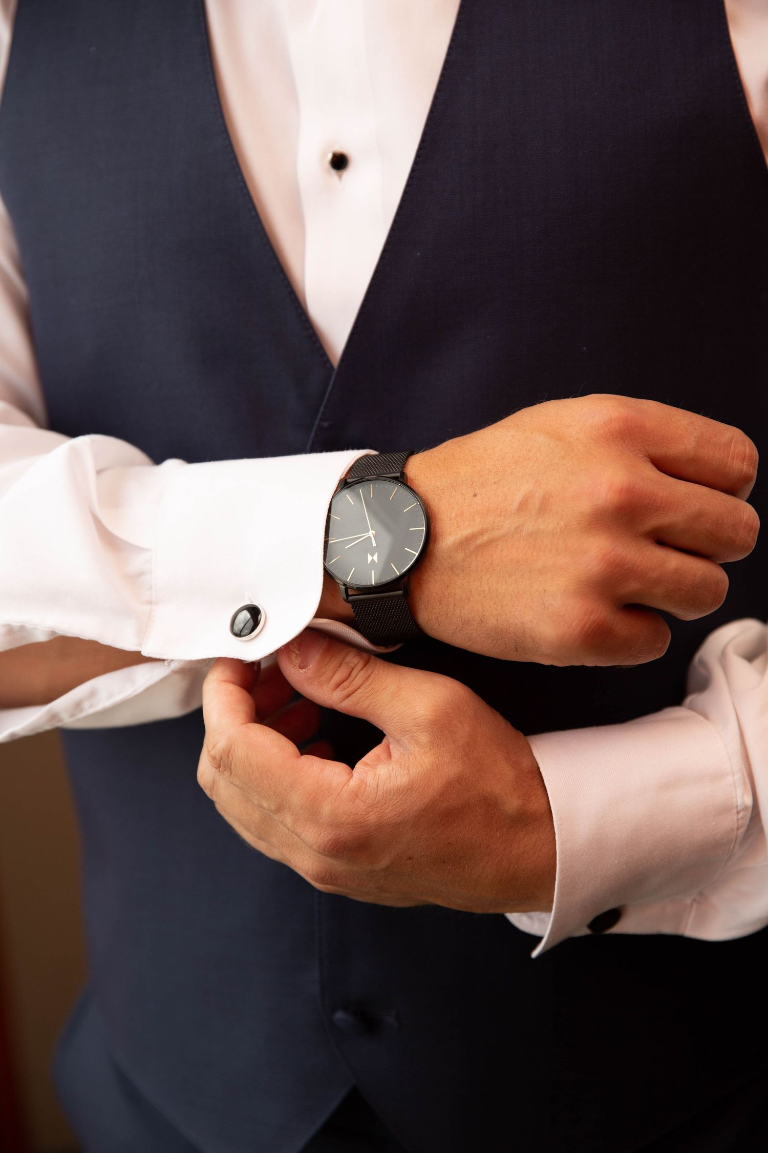 Detail shot of hands adjusting cufflinks while wearing a formal navy vest and white dress shirt.