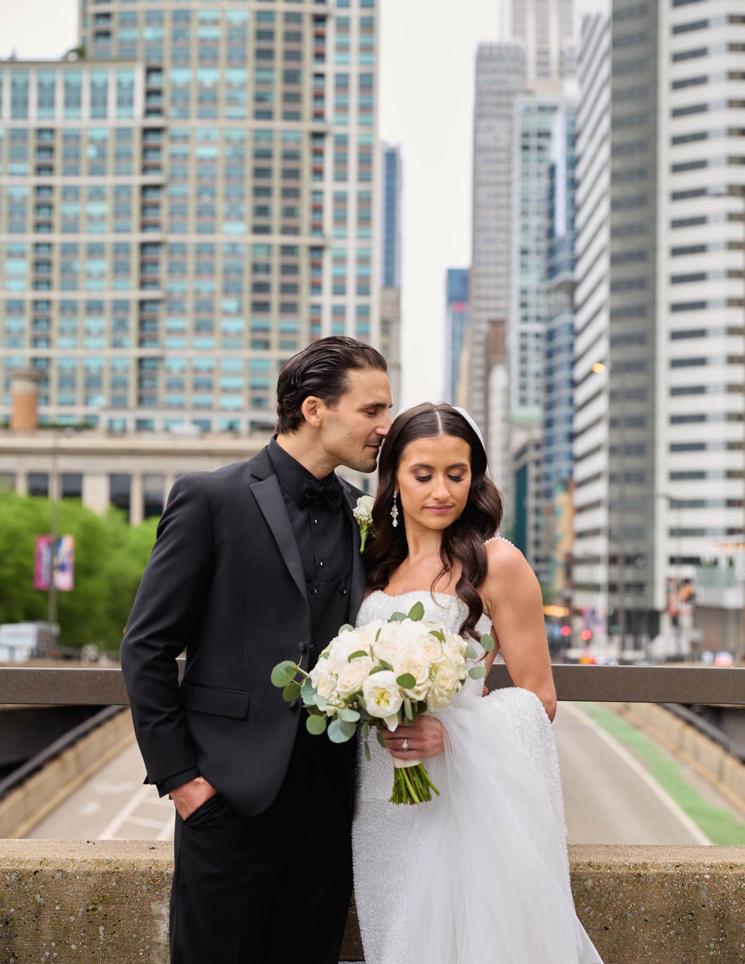 A wedding couple poses in front of Chicago skyscrapers with the bride holding a white floral bouquet.