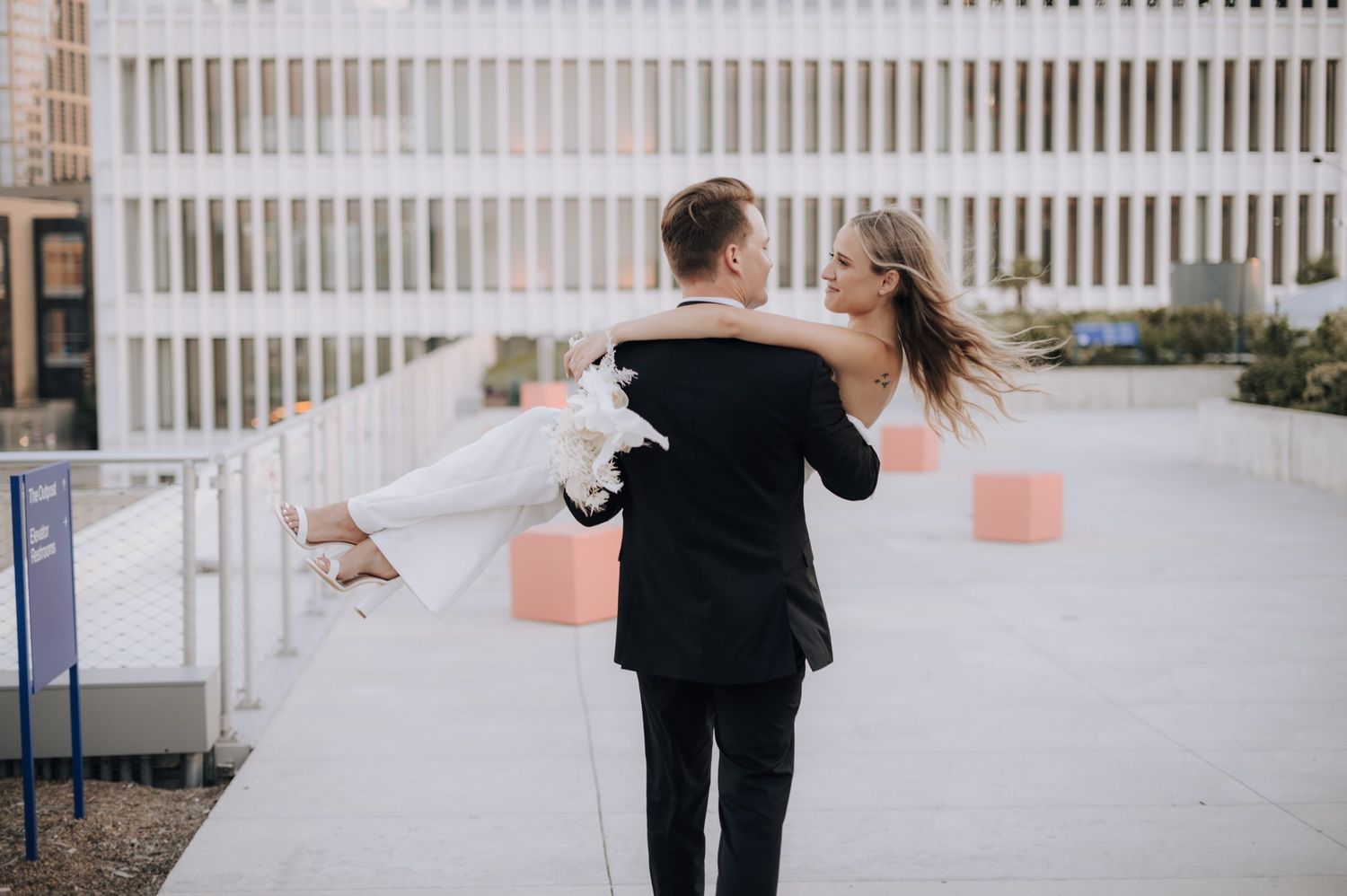 Groom carries bride in elegant white dress across an outdoor rooftop venue.