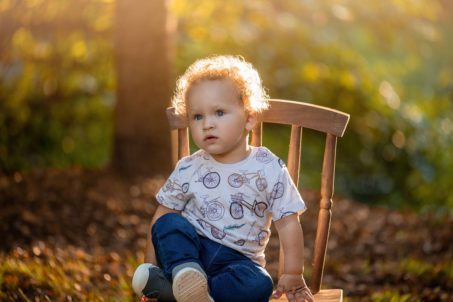 A small child sits on a wooden chair in dappled sunlight wearing a bicycle t-shirt.