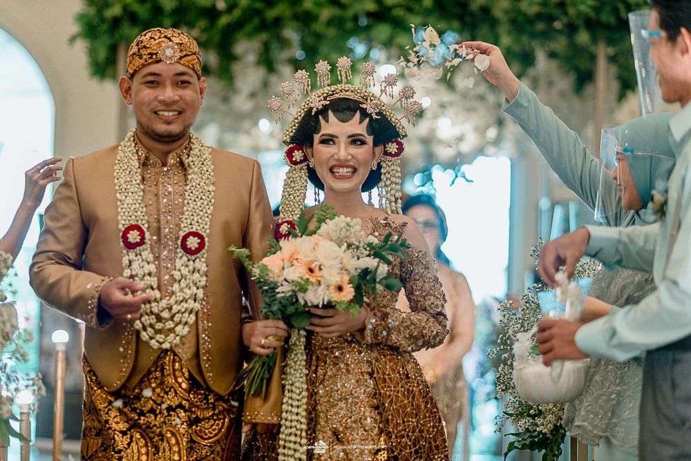Traditional Indonesian wedding ceremony with couple in golden Javanese attire and guests throwing flower petals.