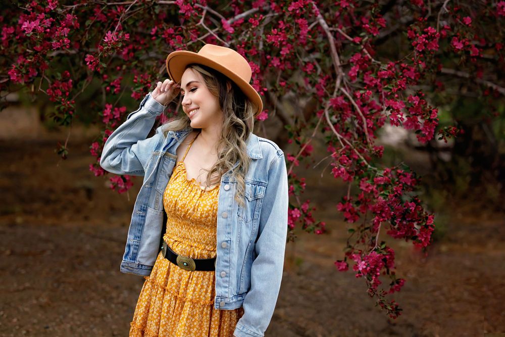 Outdoor portrait of someone in a yellow dress and denim jacket near blooming red flowers.