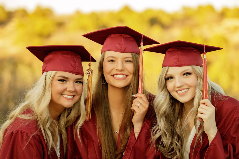 Three graduates wearing red caps and gowns celebrating against golden autumn background.