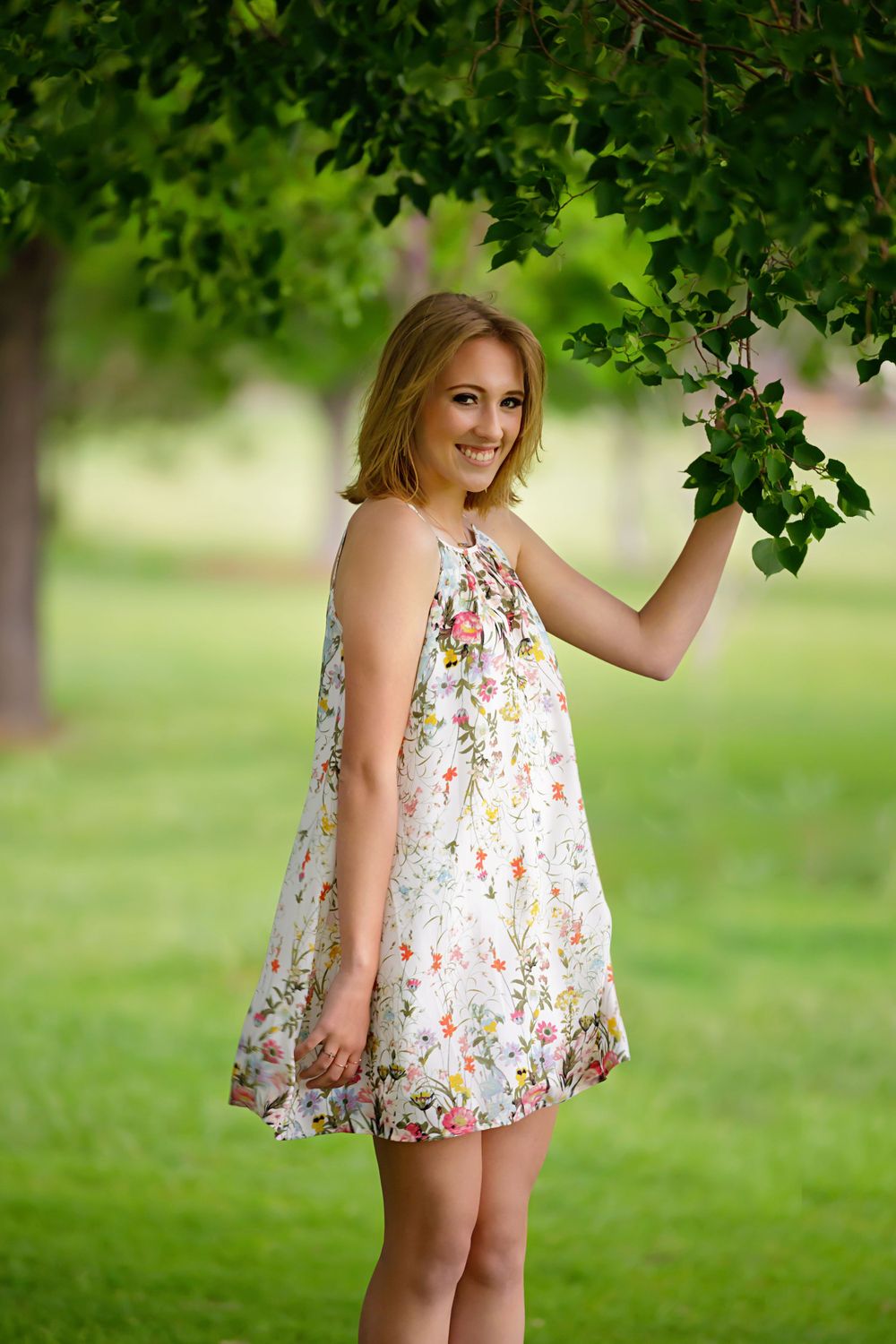 Springtime portrait of someone in a floral dress standing under green tree branches.