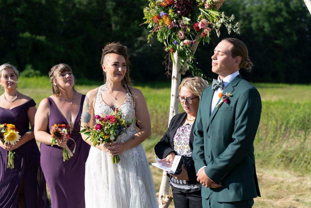 Wedding ceremony under floral arch with purple-clad attendants in summer field.