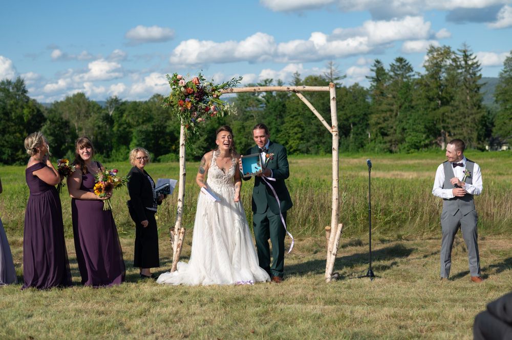 Wedding ceremony in progress with bridesmaids in purple dresses standing beside a wooden arch in a field.