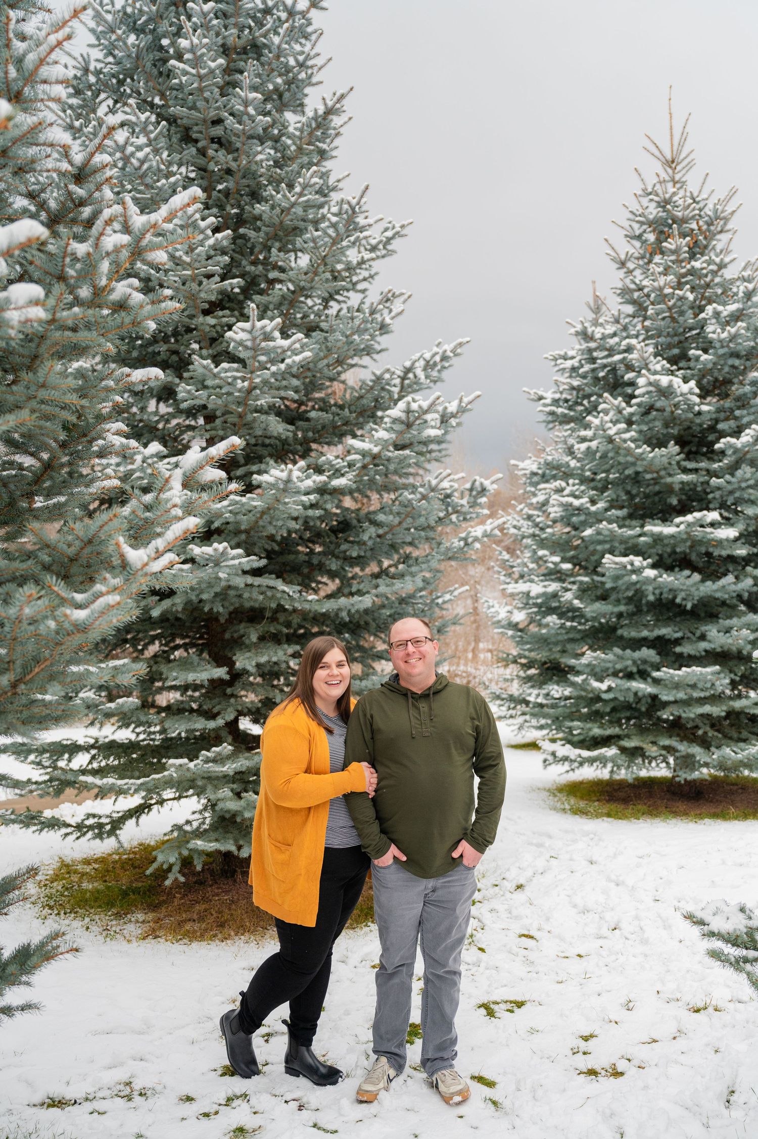 Couple standing among snow-covered pine trees wearing warm winter attire.