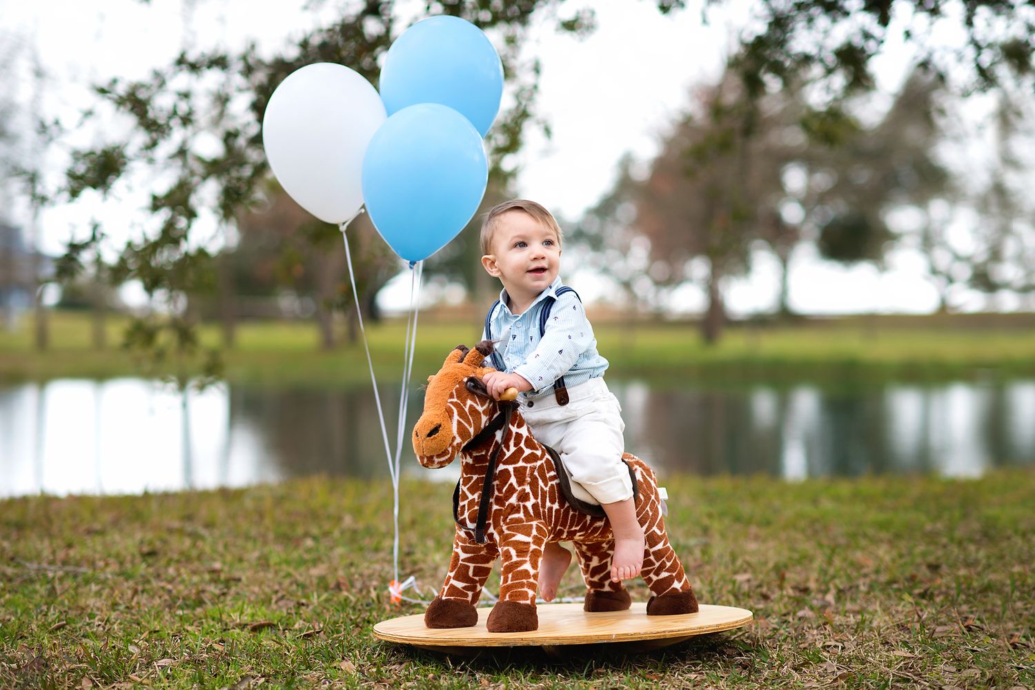 A joyful toddler rides a giraffe rocking toy surrounded by blue and white balloons outdoors.