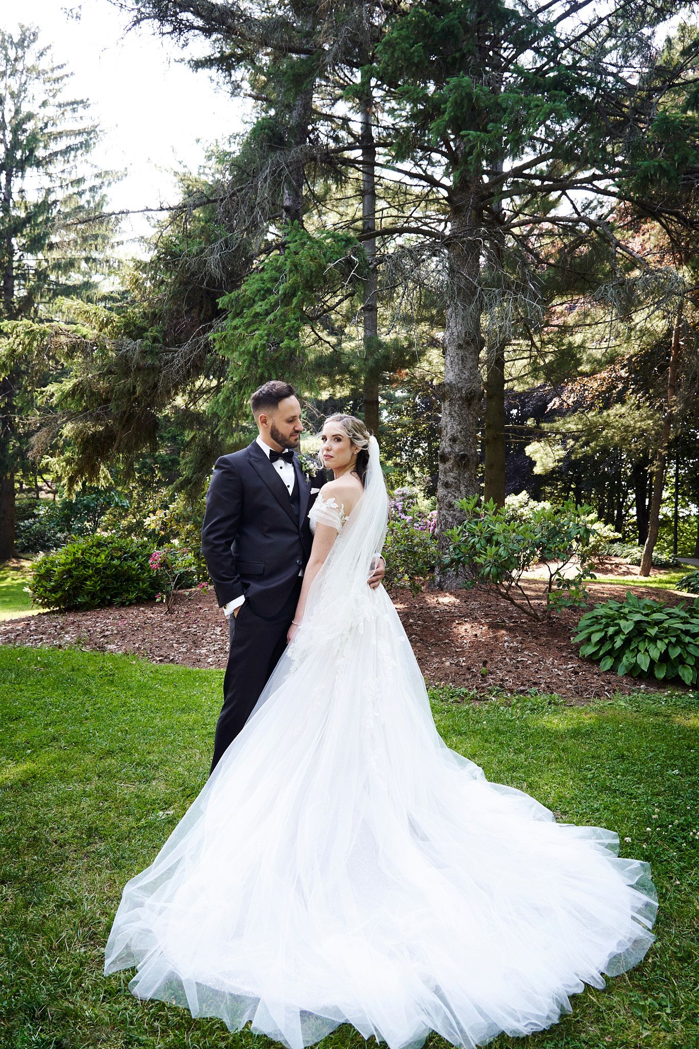 A couple poses together in a lush garden setting with tall evergreen trees and a stunning white wedding gown with flowing train.