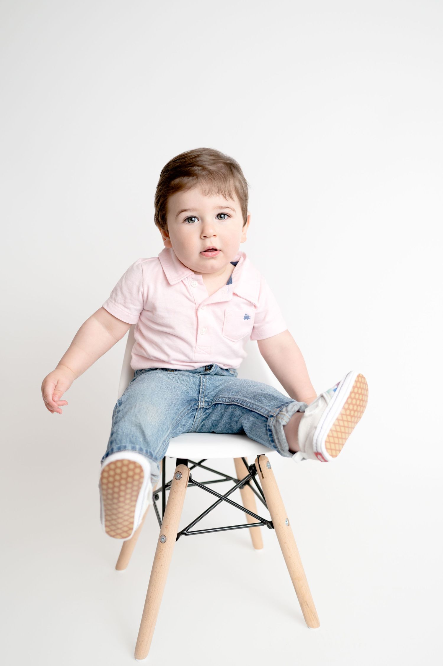 Toddler in pink shirt and jeans sits and plays on modern white chair against white background.