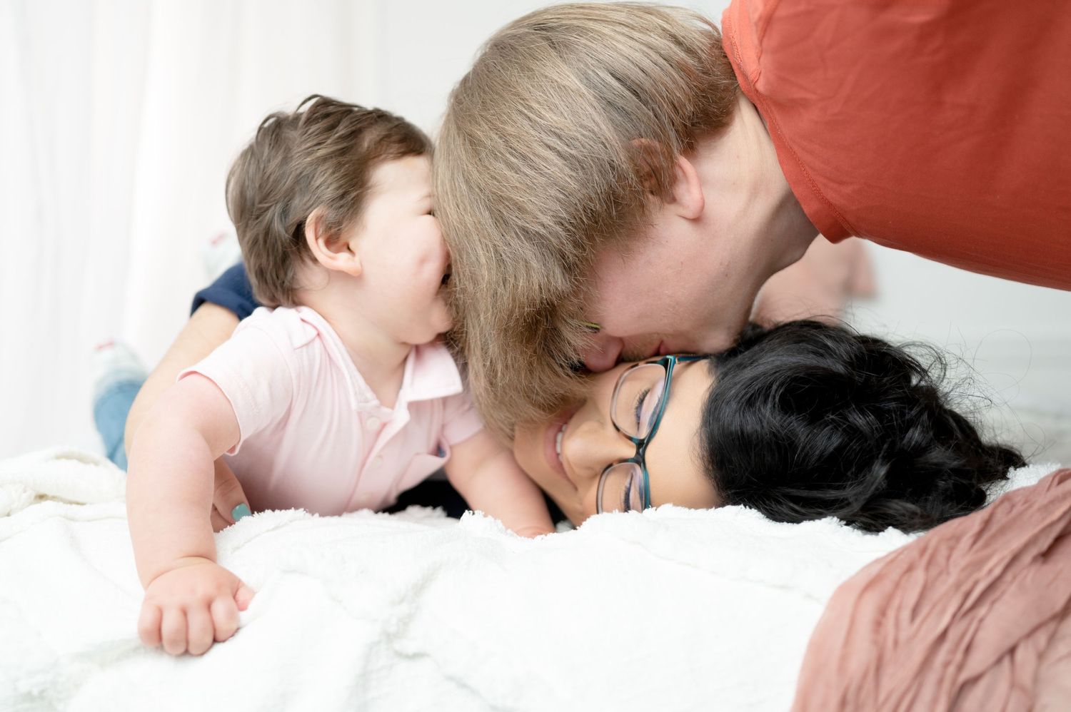 Parent and young child share a tender moment lying together on white bedding.