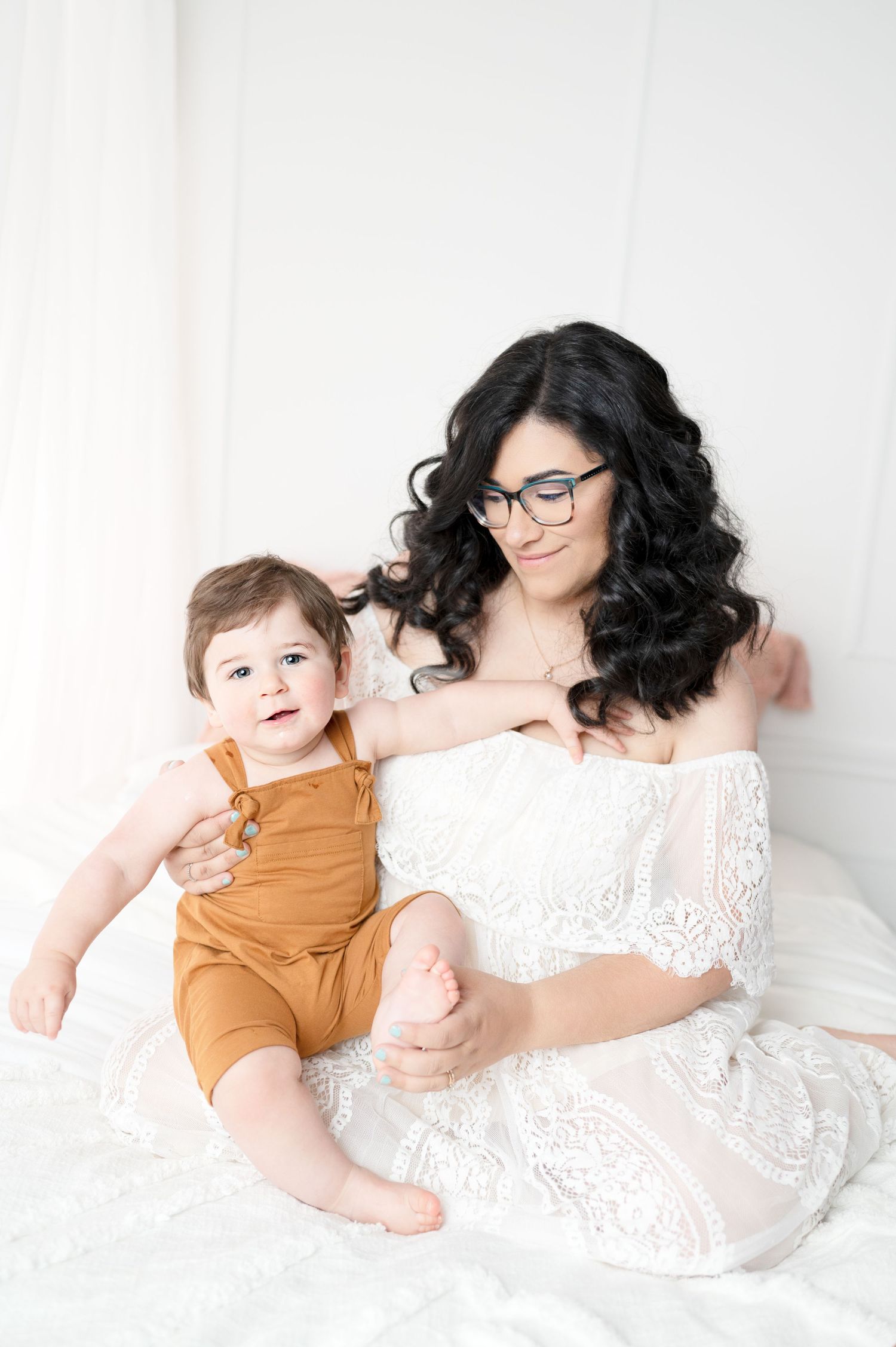 Mother in white lace dress and glasses sits with baby wearing orange overalls on white bedding.