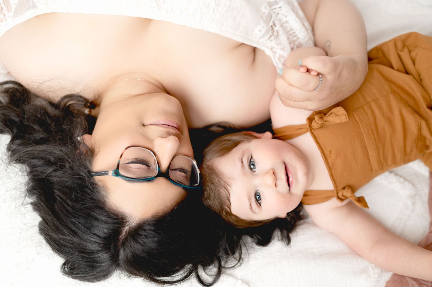 Parent and child lying on white bedding sharing joyful expressions together.