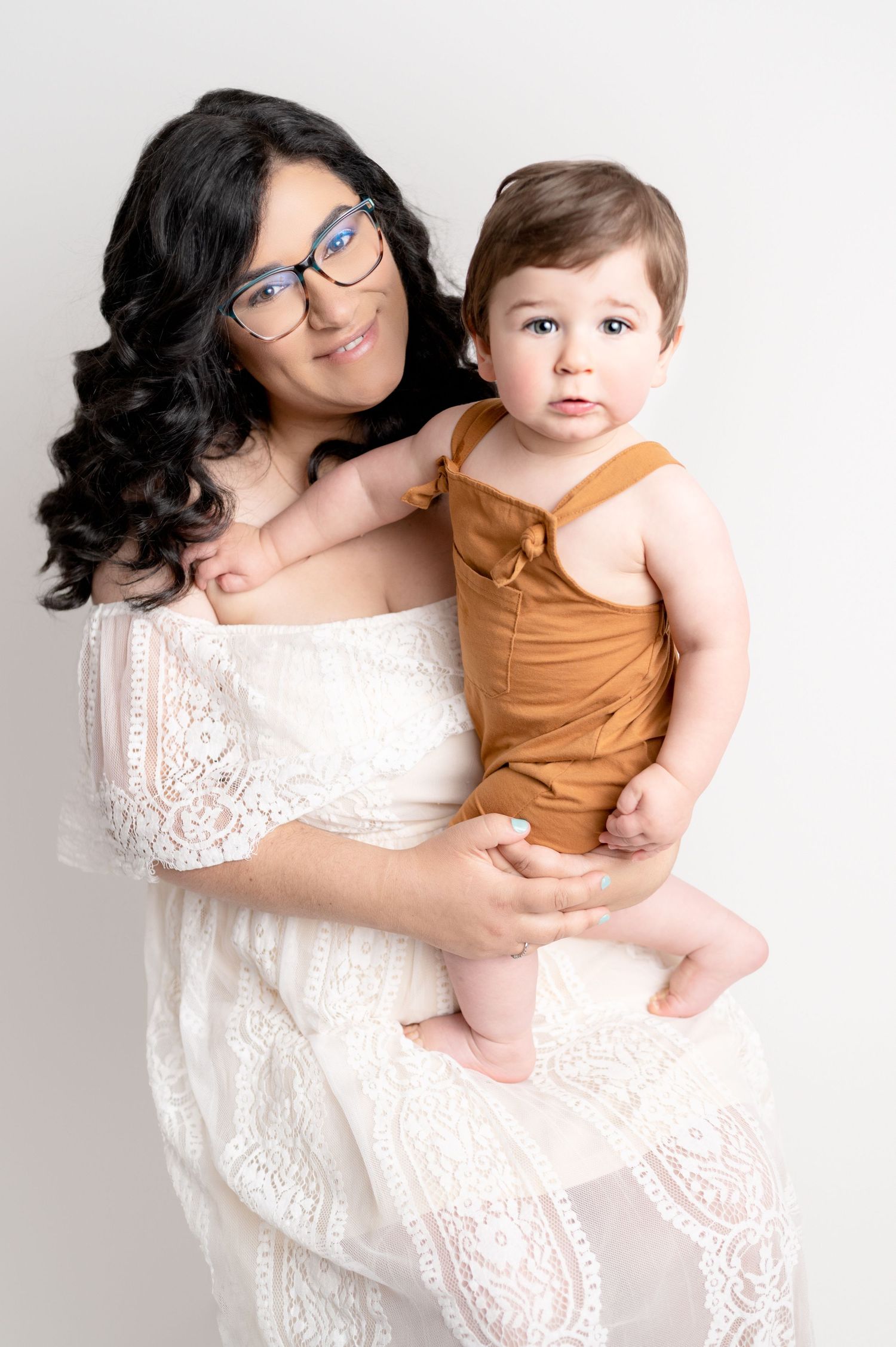 Parent in white dress holds smiling toddler in orange overalls against light background.
