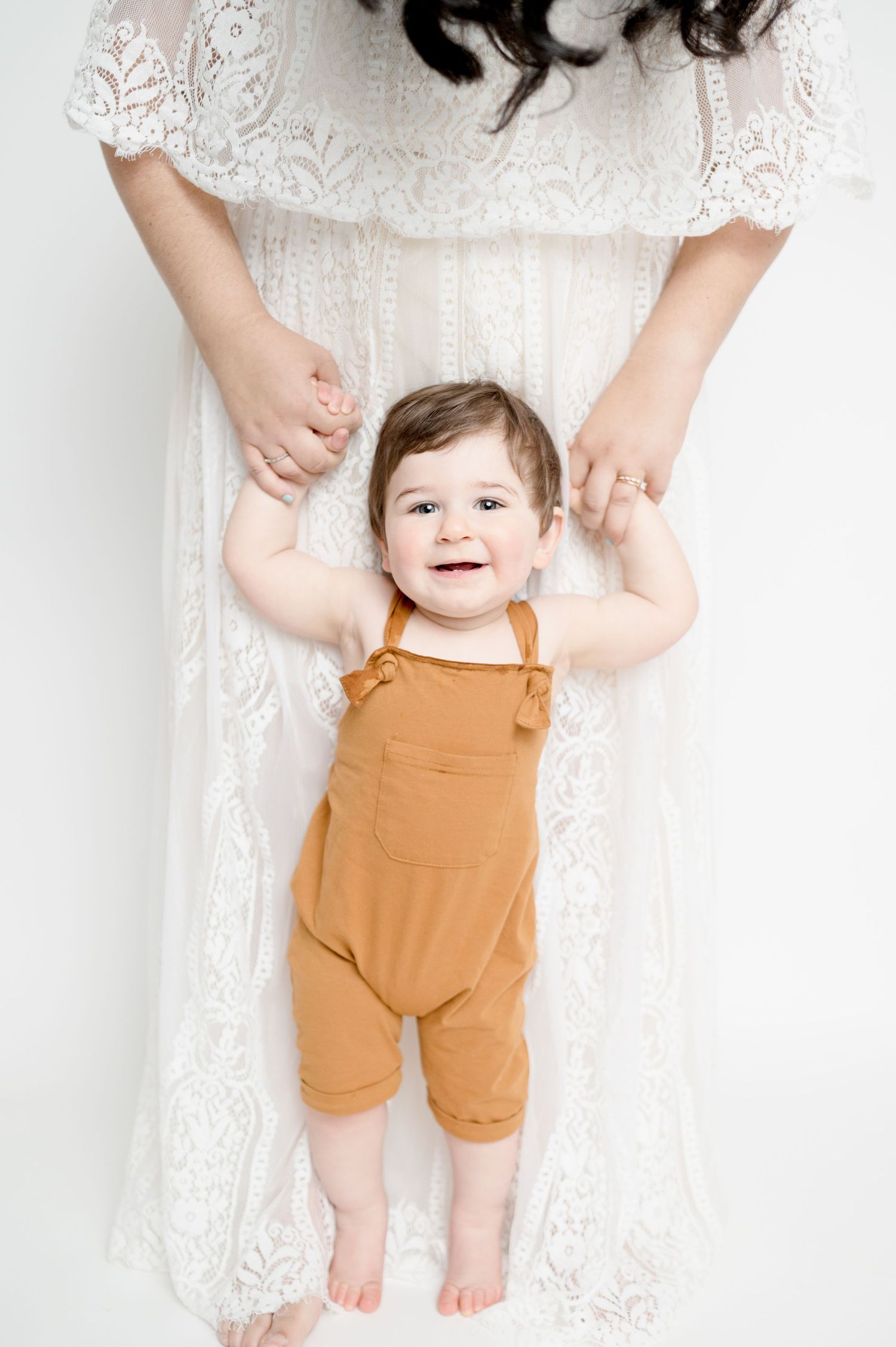 Toddler in orange overalls stands supported by parent's hands against white backdrop.