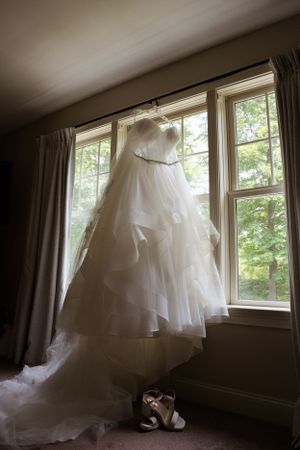 A white wedding dress hangs in front of sunlit windows with shoes placed below.