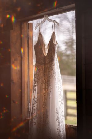 Elegant lace wedding dress hanging in window with sunlight creating rainbow prism effects.