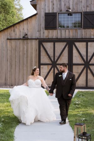 A couple in wedding attire walks along a pathway in front of a rustic barn with black trim in the Hudson Valley,NY.