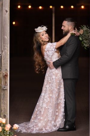 A couple shares a romantic dance pose in a lace wedding gown and suit under string lights at their barn venue.