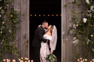 Romantic wedding couple embrace under string lights and climbing roses at rustic barn entrance.