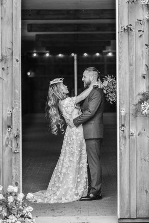 A romantic black and white wedding photo showing a couple dancing in a rustic barn doorway with string lights in the background.