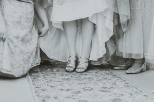 A black and white detail shot of wedding party shoes and dress hems on an ornate carpet.