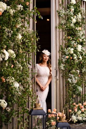 A bride in a lace dress stands framed by climbing white roses and greenery in a rustic doorway.
