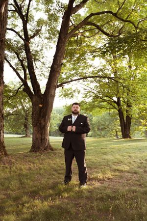 Groom in black suit stands under canopy of trees in outdoor garden setting.