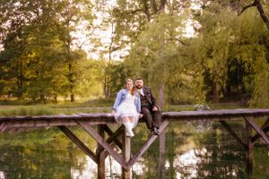 Romantic couple sitting on wooden bridge over pond during golden hour in park setting.