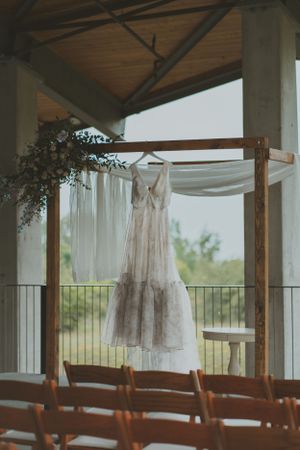 A flowing white wedding dress hangs from a rustic wooden arch with draped fabric and chairs below.