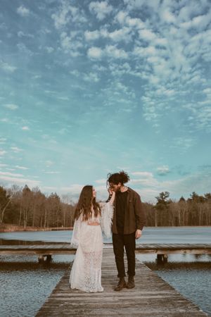 Couple in wedding attire sharing intimate moment on wooden pier with cloudy sky backdrop.