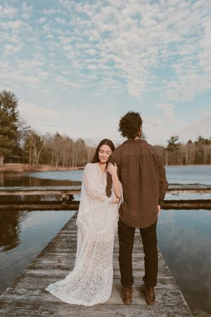 A couple stands on a wooden dock overlooking a serene lake during sunset in romantic wedding attire.