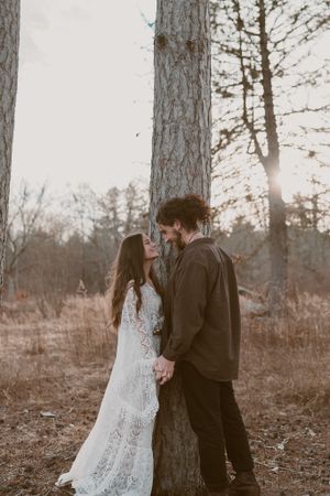 A romantic moment between a couple in a lace gown and brown shirt against tall pine trees at dusk.