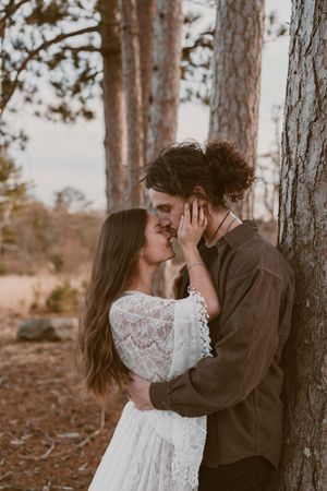 An intimate embrace between two people in wedding attire among tall pine trees.