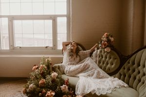 Lace-clad figure reclining on a tufted velvet sofa with floral arrangements.