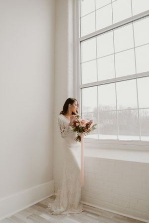 A bride in a white lace gown stands by a large window holding a pink and white floral bouquet.
