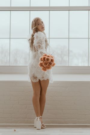 A bride in a short lace dress holding a peach rose bouquet poses near a bright window.