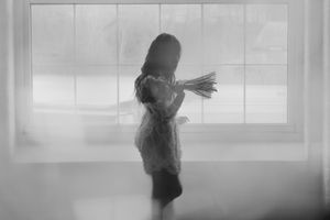 A silhouetted figure holds flowers while standing against a bright window backdrop.