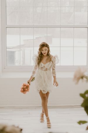 A bride in a short white lace dress twirls while holding a peach-colored bouquet.