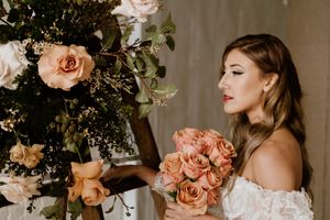 Person in off-shoulder dress holding peach roses next to elegant floral arrangement.