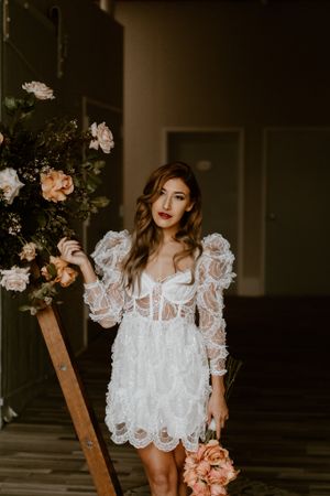 A bride in a white mini dress poses next to a floral arrangement on a wooden ladder.