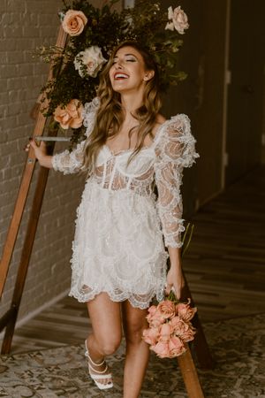 Short white lace dress with long sleeves and pink roses in a romantic vintage-inspired wedding photoshoot.