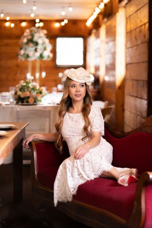 Bride in vintage-inspired lace dress poses on red velvet couch in rustic wood venue.