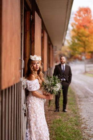 Bride in lace dress holds bouquet while groom waits in background during fall photoshoot.