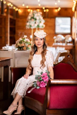 Elegant bride with pink fascinator sitting on red velvet sofa holding floral bouquet.