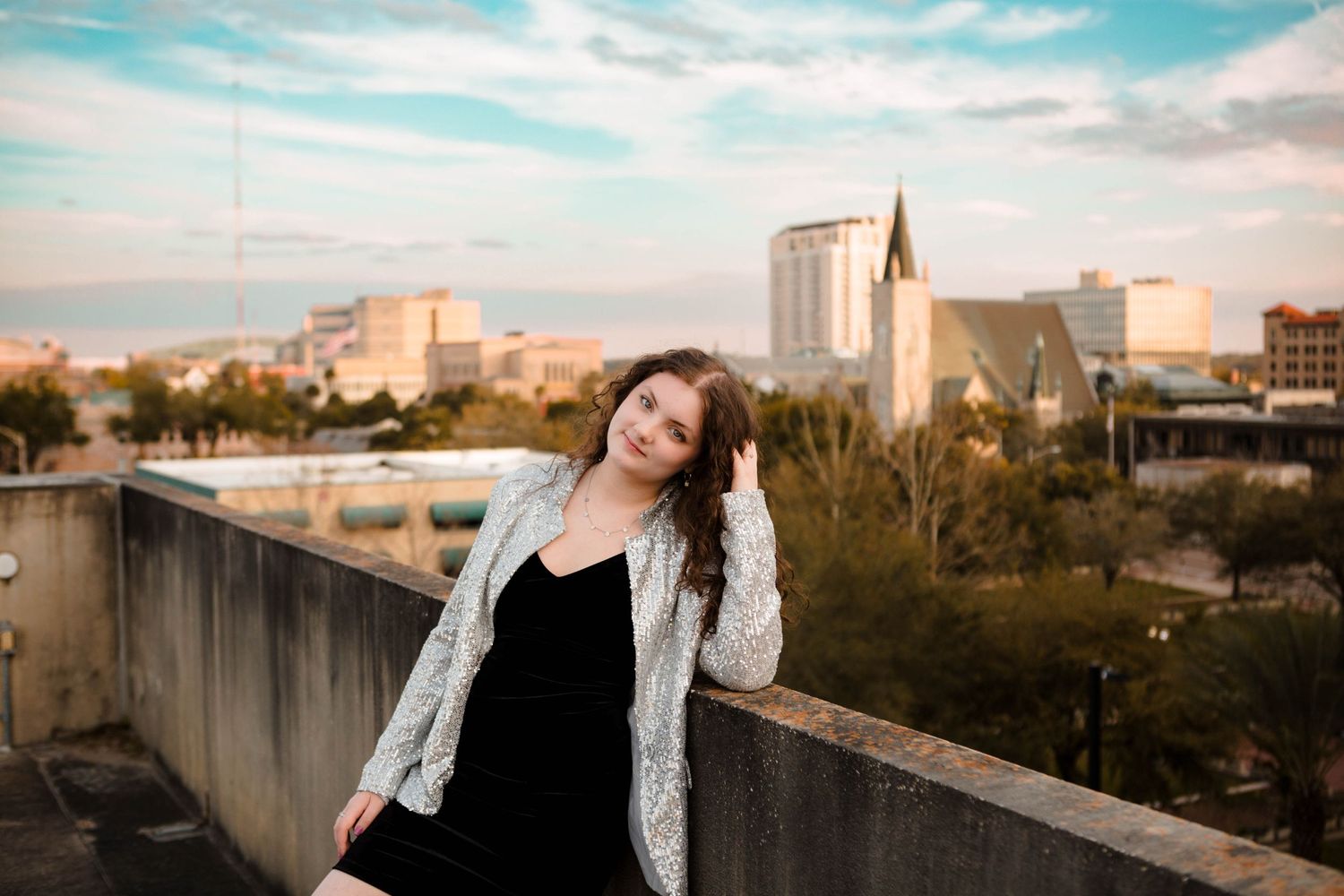 A woman in black dress and white cardigan poses on an urban rooftop with a cityscape backdrop.