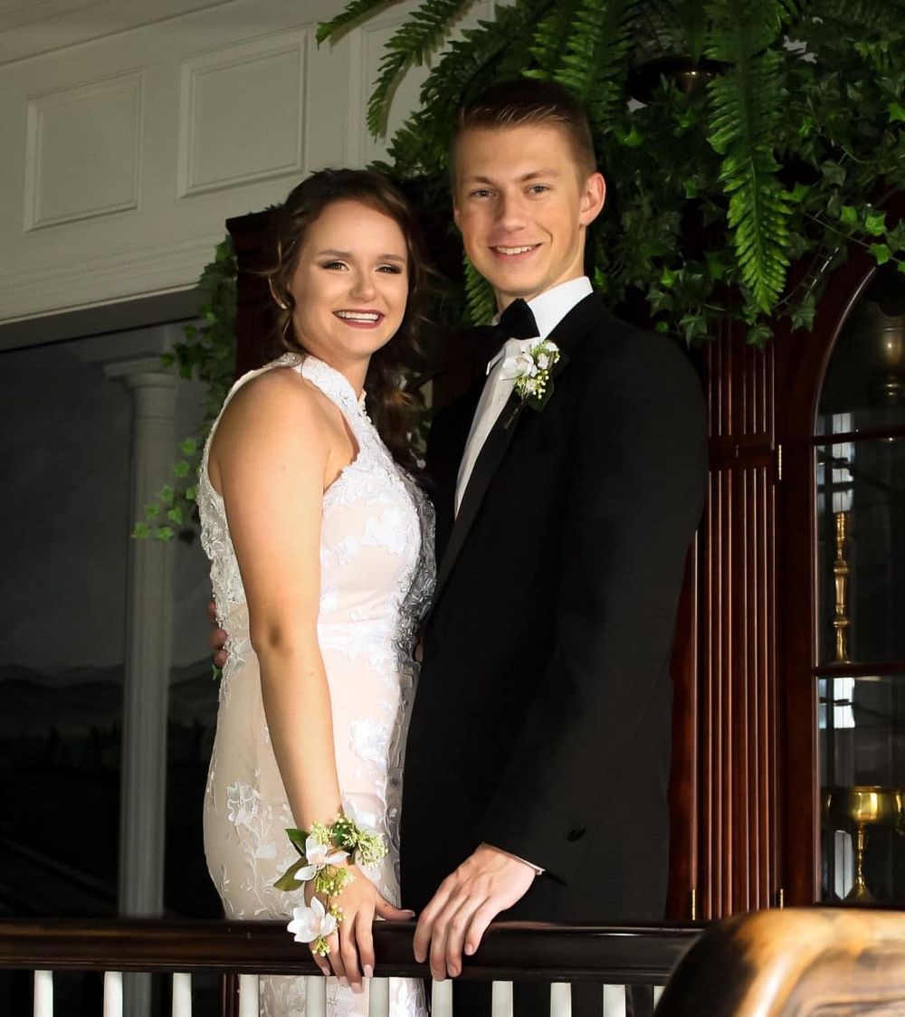A formal couple poses together in front of decorative woodwork and greenery.
