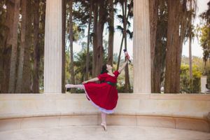 Ballerina in red dress performing arabesque pose between classical stone columns outdoors.