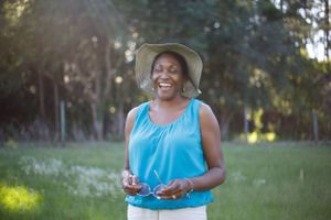 Happy senior Black woman in blue top and hat laughing outdoors in sunny park.