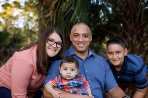 Smiling family of four with baby posing together outdoors among tropical trees.